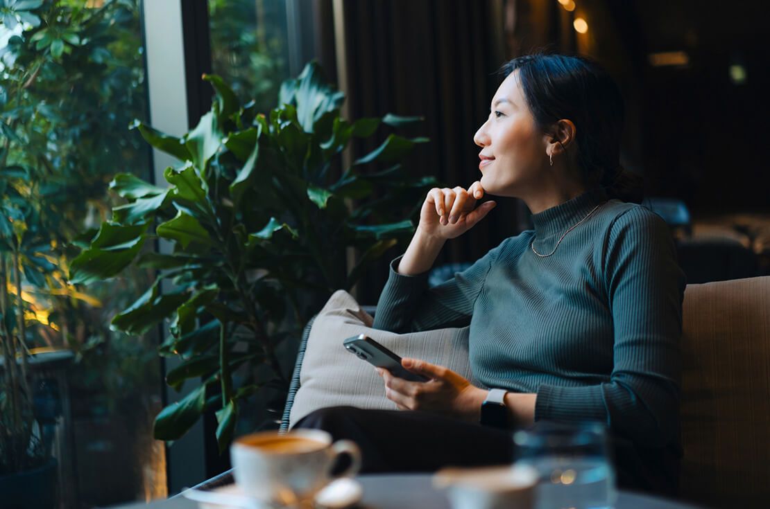 Confident young Asian woman with smartphone looking out through window while sitting in a cafe having coffee. Making a personal financial plans and investment decisions. Wealth management. Business, banking, finance and investment concept